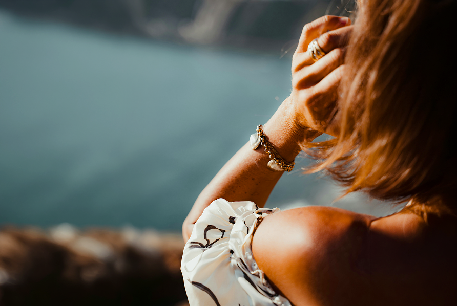 Nainen veden ��rell� ilta-auringossa olan yli takaap�in kuvattuna k�si korvanjuuressa, rannekoru ranteesssa.