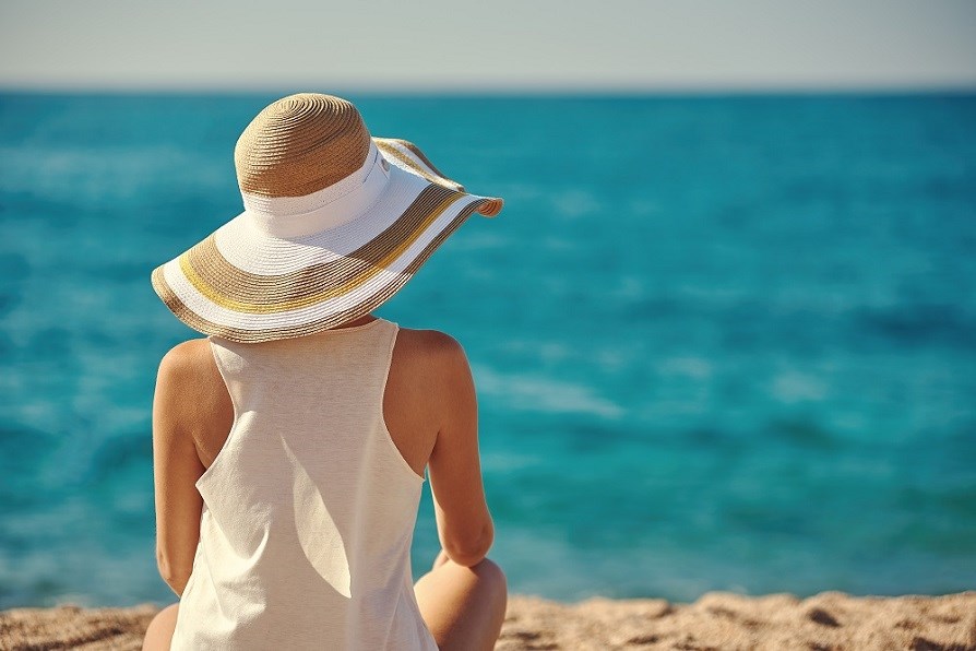 Nainen hattu pss ja toppi ylln istuu rannalla ja katsoo merelle.