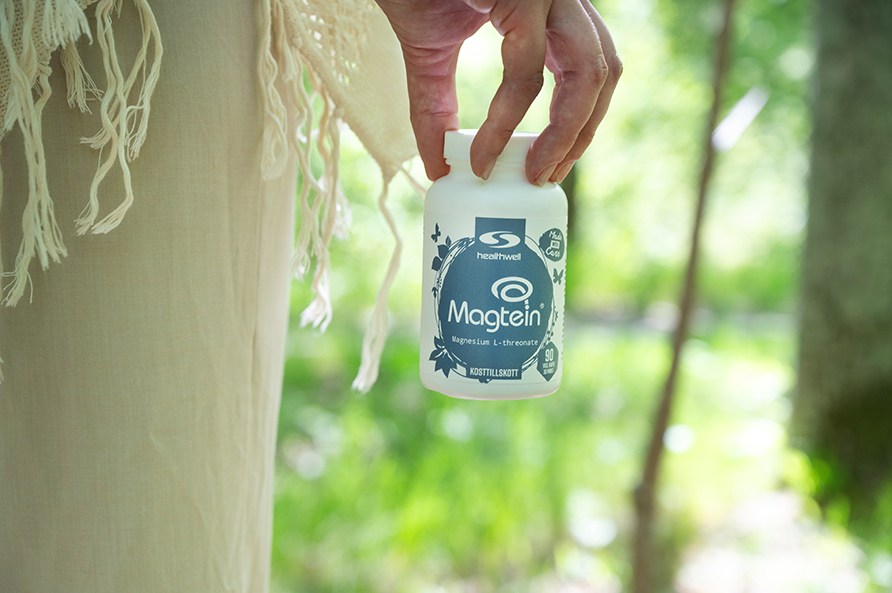 Naisen sormet roikottavat Healthwell Magnesium Magtein -purkkia mets�ist� taustaa vasten.