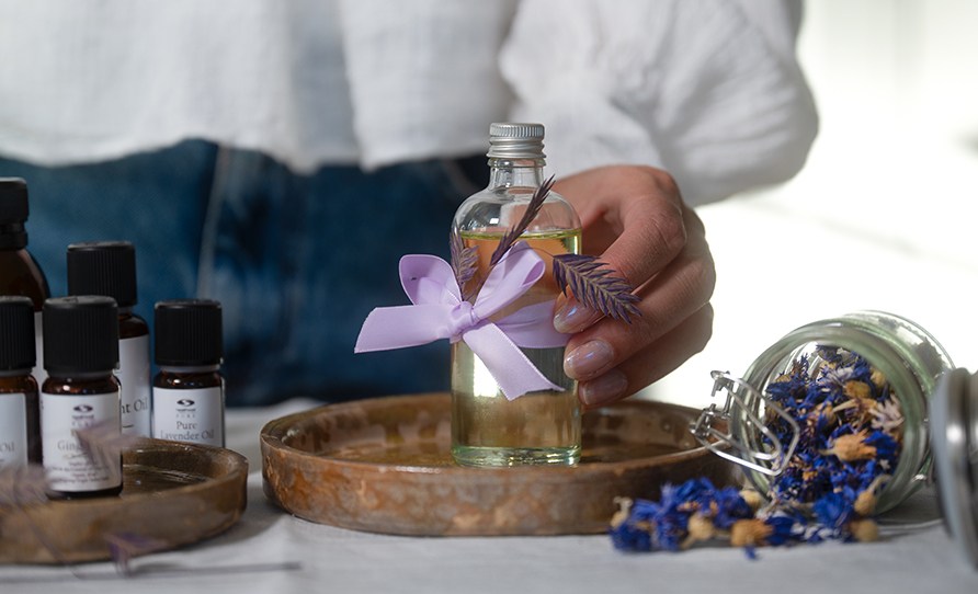 Nainen ottamassa lasista hierontaljypulloa keraamiselta tarjottimelta. Pydll mys eteerisi ljyj ja kuivattuja kukkia purkissa.