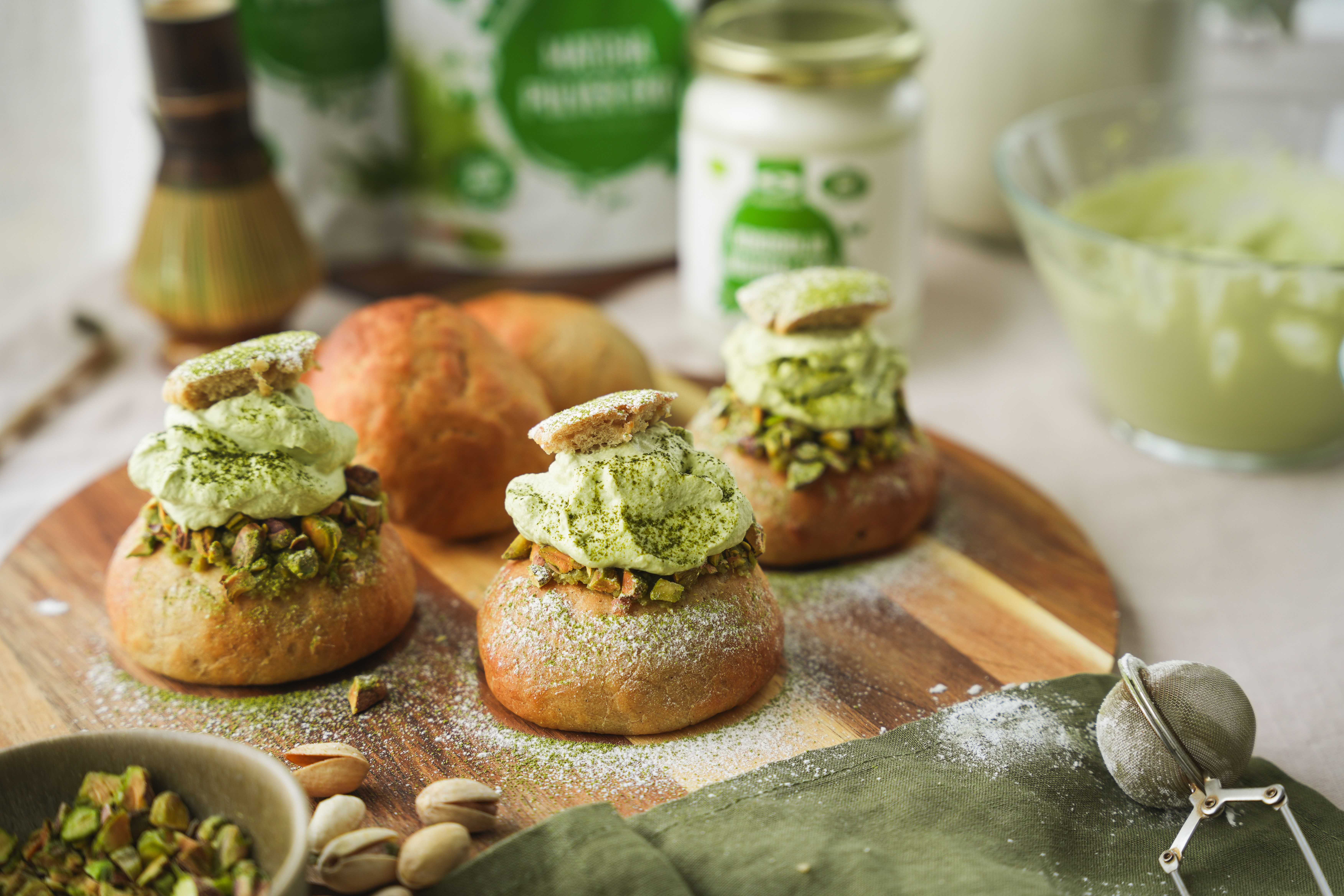 Herkullisia matchapistaasi-laskiaispullia, kauniin vihre�ll� kermalla puutarjottimen p��ll�, ymp�rill� tarvittavia aineksia.