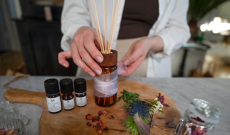 Nainen ottamassa lasista puutikuilla varustettua tuoksupulloa puiselta leikkuulaudalta, jolla myös eteerisiä öljyjä ja kuivakukkia. Nainen ottamassa lasista puutikuilla varustettua tuoksupulloa puiselta leikkuulaudalta, jolla myös eteerisiä öljyjä ja kuivakukkia.